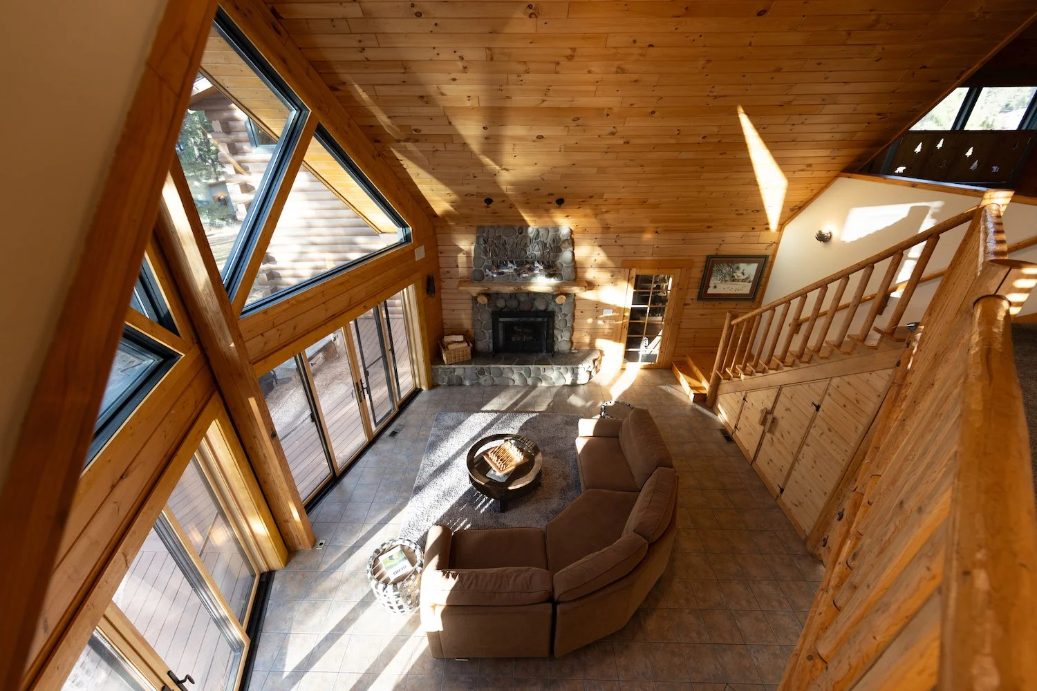 A spacious living room with wood-paneled walls and ceiling, large windows, a stone fireplace, a beige sectional sofa, a coffee table, and a wooden staircase leading to a second floor.