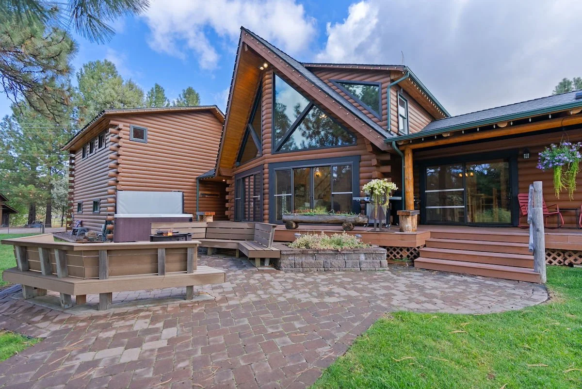 A modern log cabin home with large glass windows and a wooden deck in a wooded area, featuring outdoor seating, plants, and a brick-paved patio.