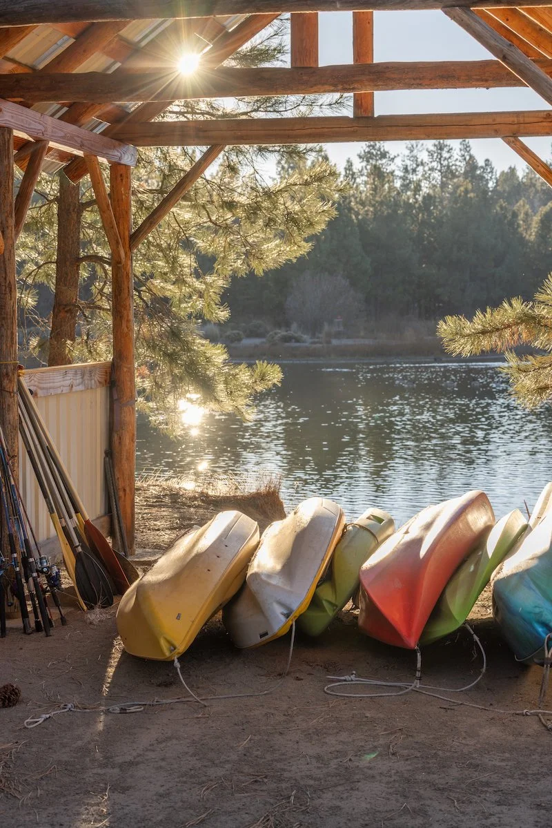 Colorful kayaks turned upside down on a beach near a lake, with fishing rods leaning against a wooden and metal structure, and trees in the background.