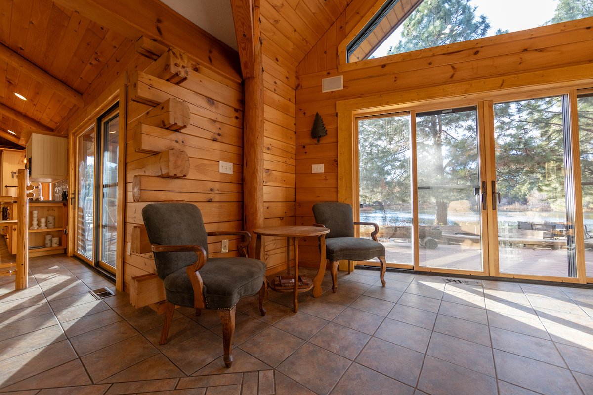 Interior of a wooden cabin with two upholstered chairs, a small round wooden table, large glass sliding doors, and a view of trees outside.