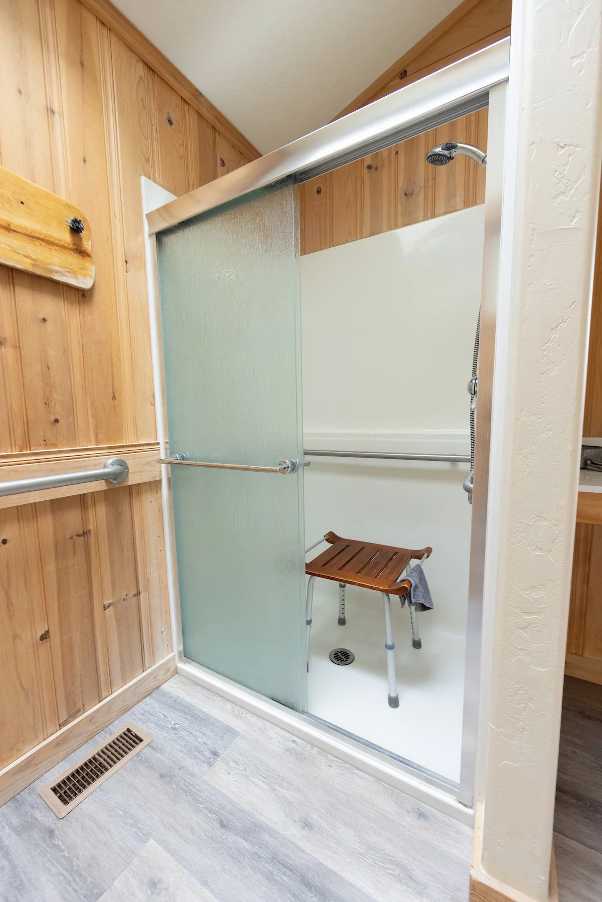 Shower stall with frosted glass sliding door, wooden bench, and handheld showerhead, framed by wood-paneled walls.