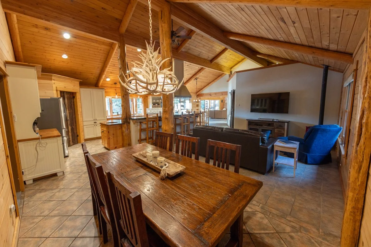 Open-concept living and dining area with wooden accents, vaulted ceiling, and modern kitchen in the background. There is a large wooden dining table with chairs and a decorative antler chandelier hanging above it. The living area has a sofa, an armch