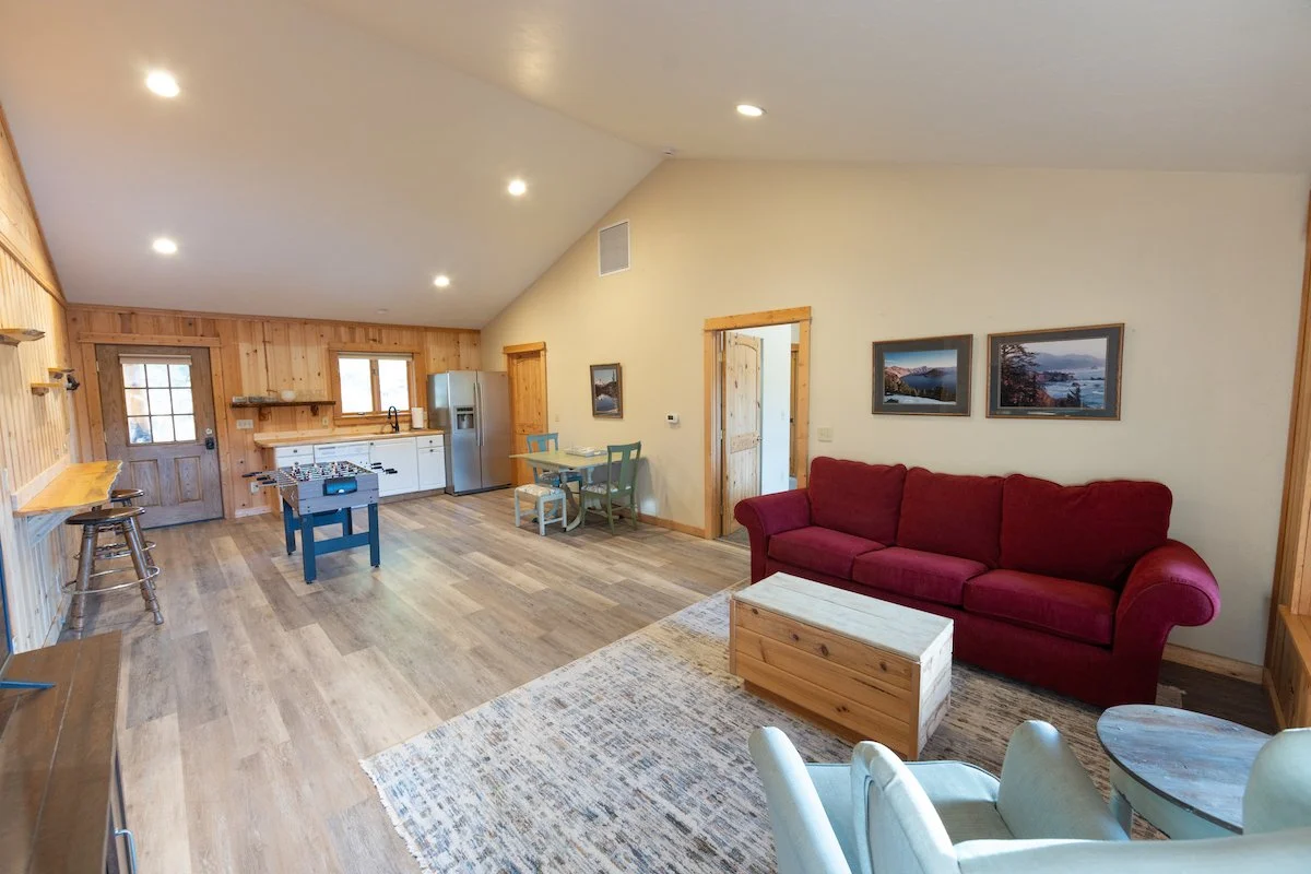 Open living space with wood-paneled walls, a red sofa, a small dining table with chairs, and a kitchenette with white cabinets and a refrigerator.