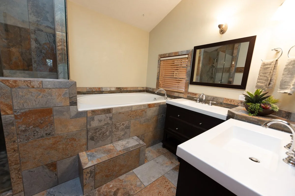 A bathroom with a bathtub, a vanity with two sinks, a large mirror, a window with wooden blinds, and beige walls with towel hooks holding two towels.