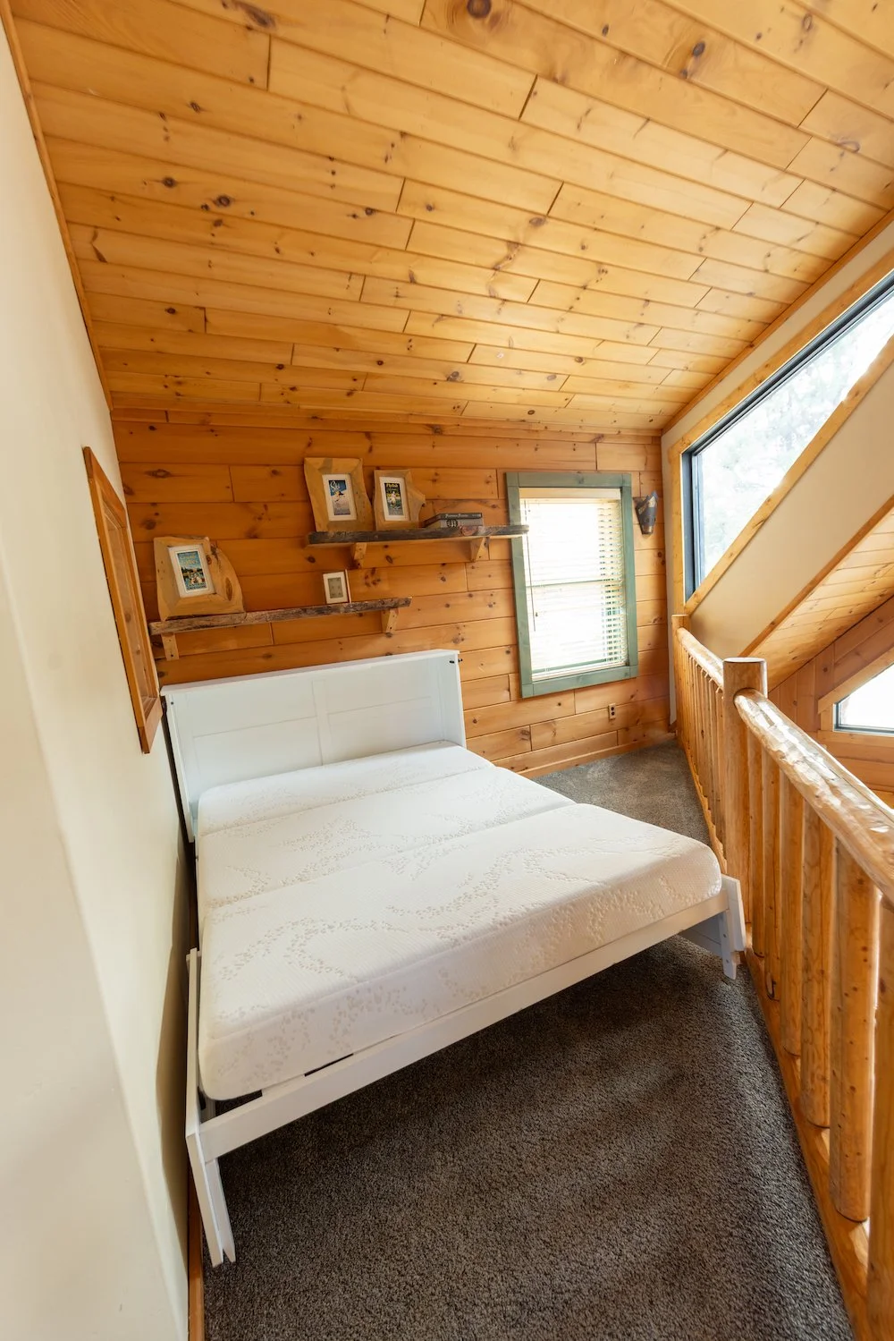 A bedroom with wooden walls and ceiling, a white bed, wooden shelves with picture frames, a small window with blinds, and a large angled window with a wooden railing.