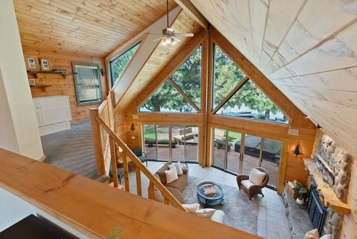 A spacious living room with a high vaulted ceiling made of wood, large triangular windows, a stone fireplace, and patio doors leading to an outdoor deck with trees outside.