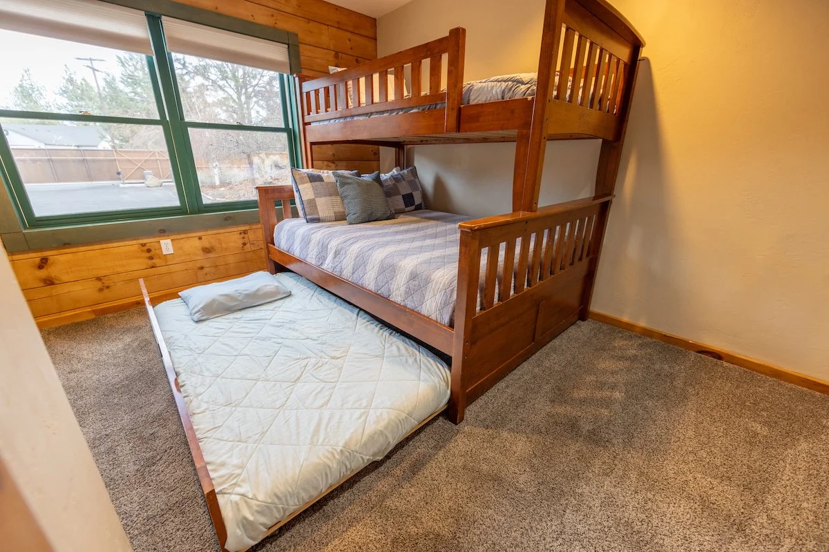 A bedroom with a wooden bunk bed and a trundle bed, a window with a view of trees and a fence outside, and a carpeted floor.
