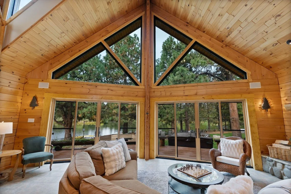 Living room with high vaulted wooden ceiling, large triangular windows showing trees and water outside, sliding glass doors leading to an outdoor deck, cozy furniture including a beige sofa, a brown armchair with a white pillow, a teal armchair, a co