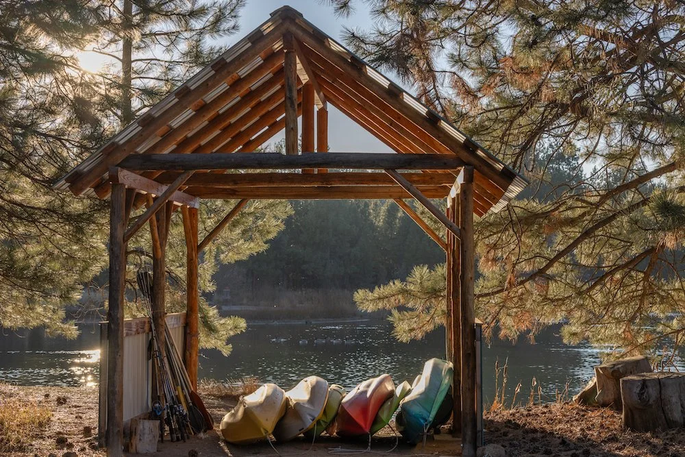 A rustic boathouse with a sloped roof sits by a lake, surrounded by pine trees. Several canoes are lined up on the ground inside the boathouse.