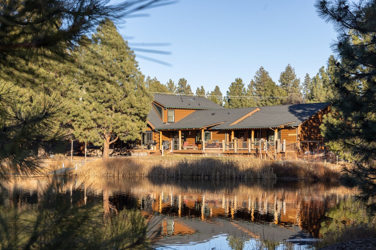 A large wooden house with a gray roof beside a pond, with trees and shrubs around it, in a rural setting.