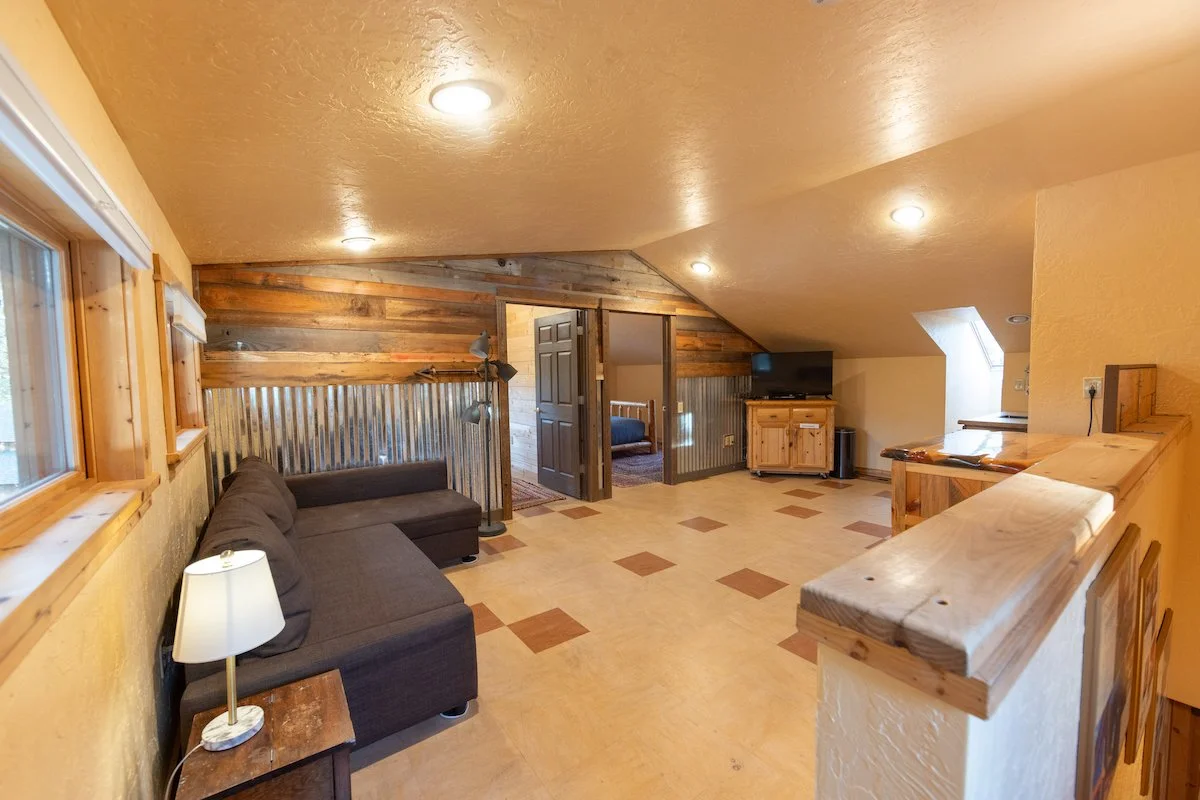 A cozy living room with wood-paneled walls, a dark gray sectional sofa, a small wooden end table with a lamp, a floor lamp with multiple adjustable arms, a flat-screen TV on a wooden cabinet, and a small window with a ceiling light, overlooking a bed