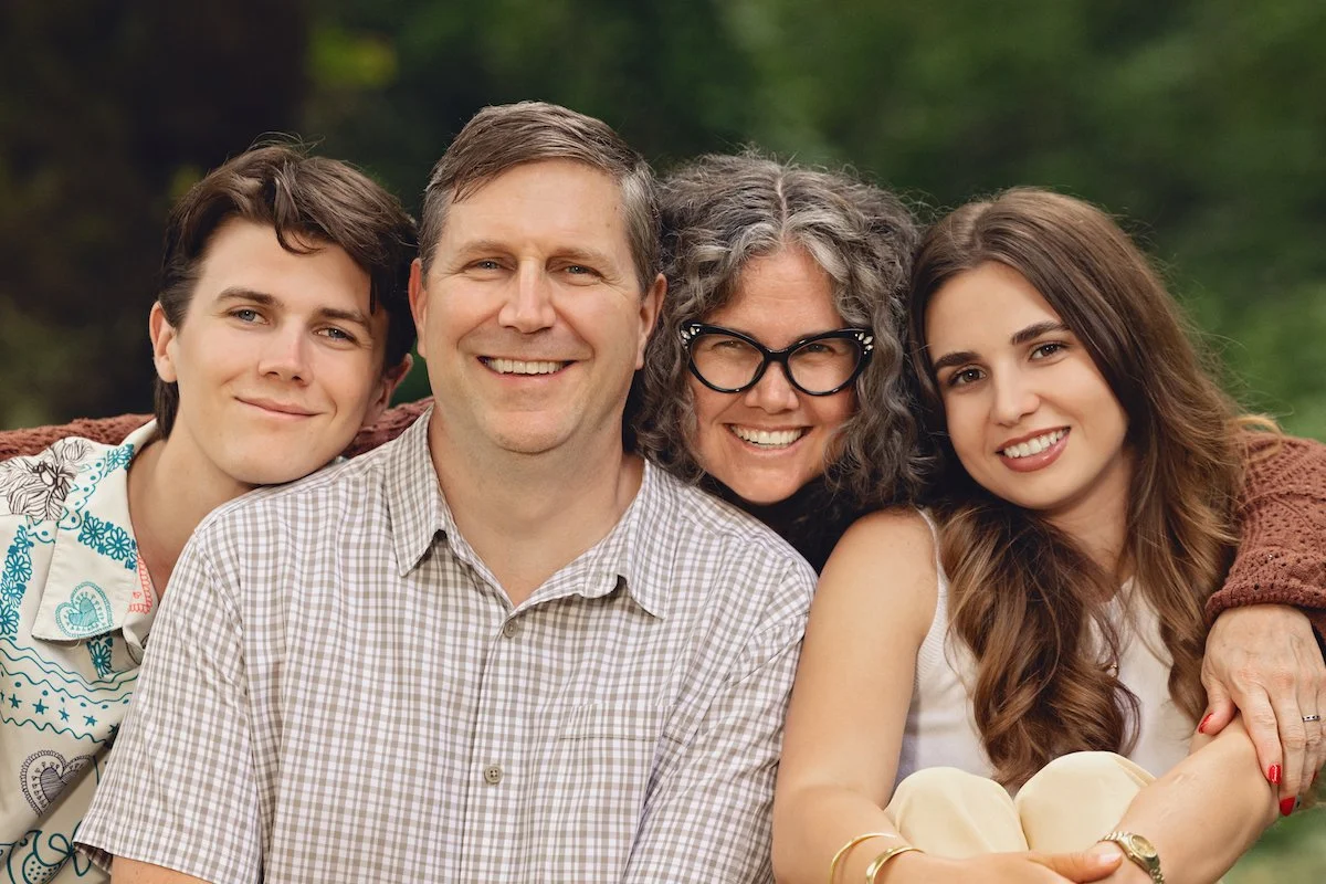 A family of four smiling outdoors, sitting closely together with a blurred green background.