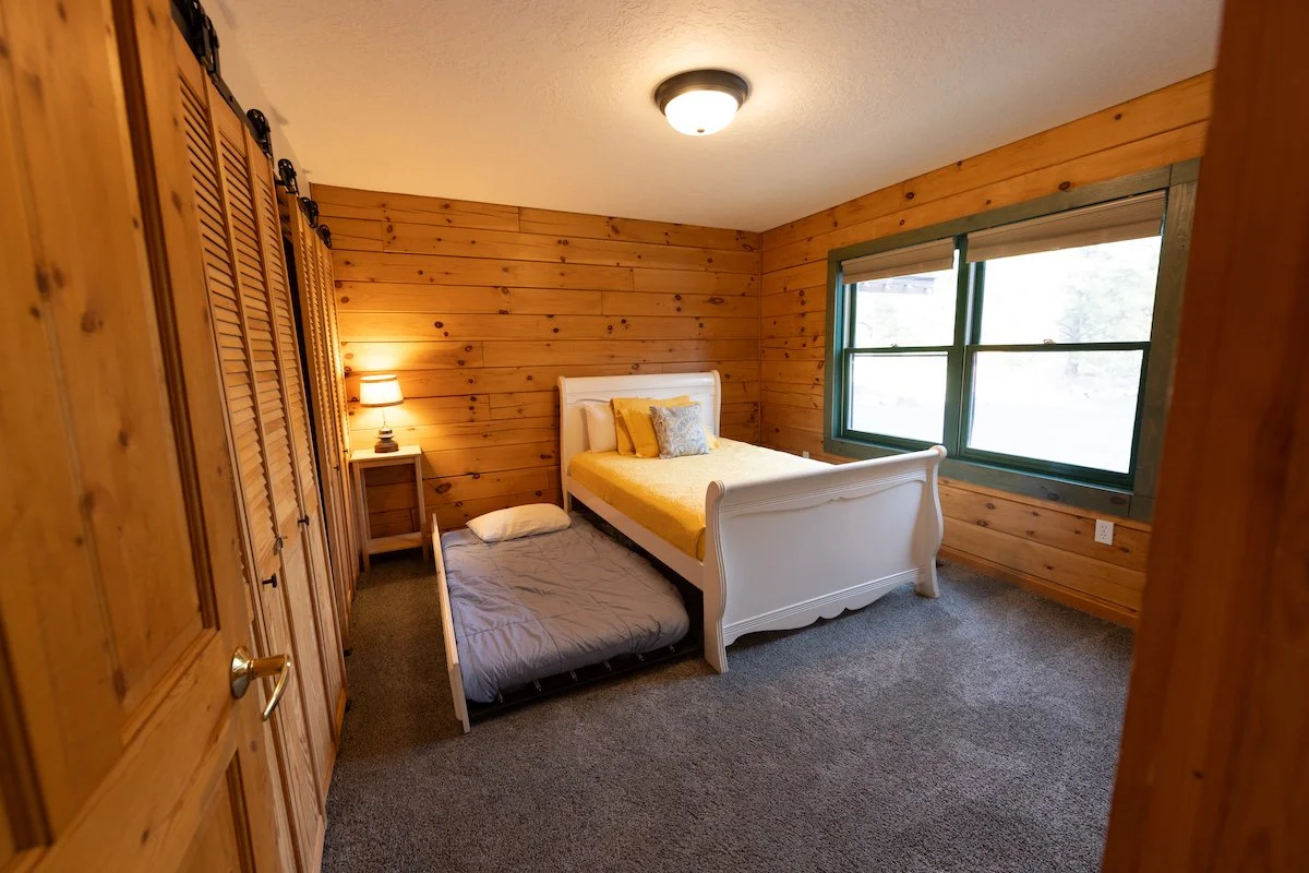 A bedroom with wood-paneled walls, a white bed with yellow and gray pillows, a nightstand with a lamp, a wooden wardrobe, and a window with green trim.