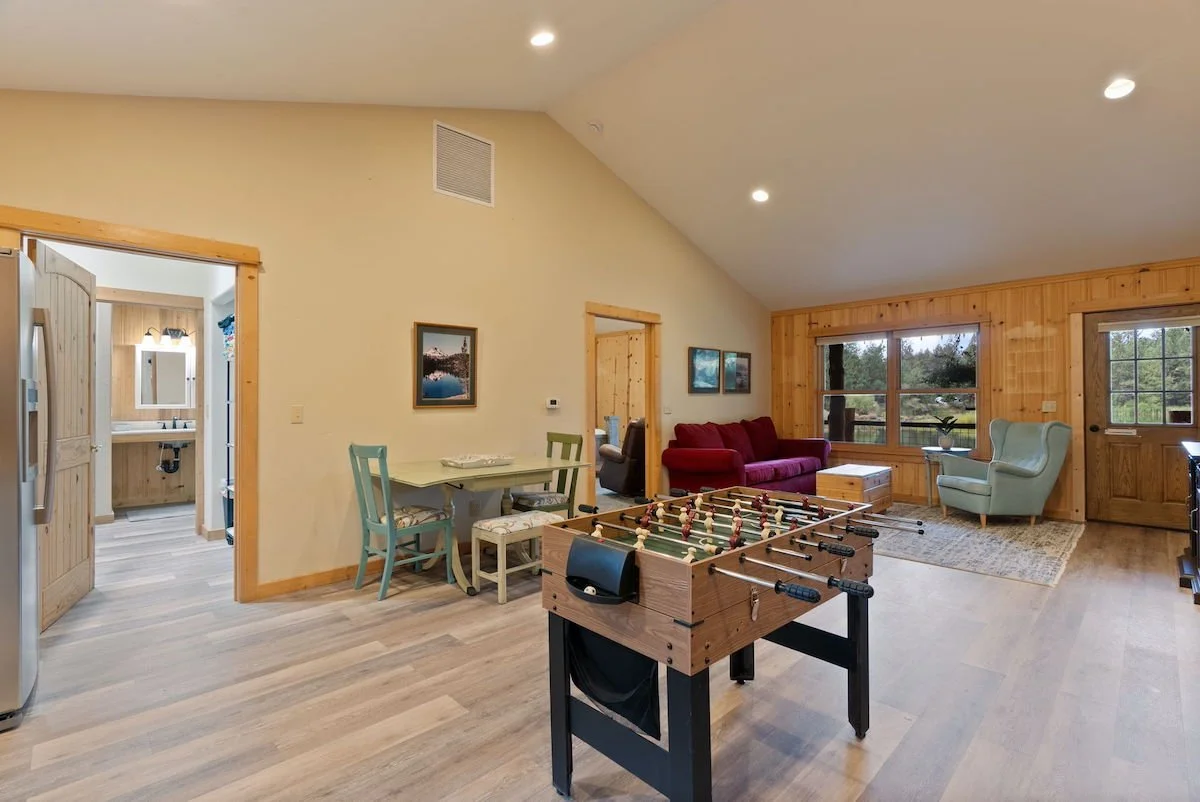 Living room with foosball table, red sofa, light blue armchair, wooden walls and floors, large windows, and a doorway leading to a bathroom.