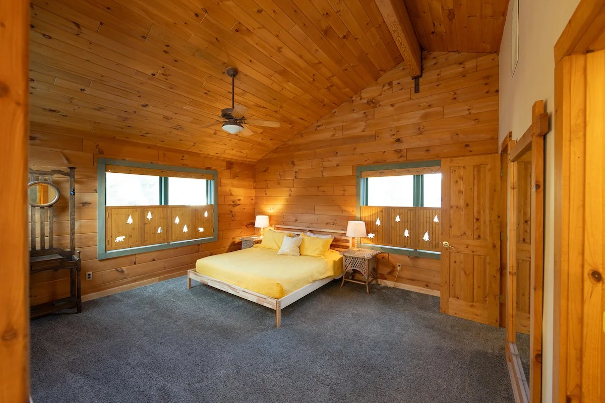 A bedroom with wooden walls, ceiling, and door, featuring a bed with yellow bedding, two nightstands with lamps, two windows with forest scenes, and a ceiling fan.