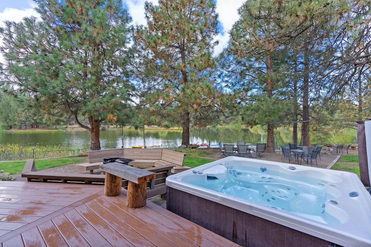 Outdoor patio area with a hot tub, wooden bench, trees, and lake view with chairs and small boats