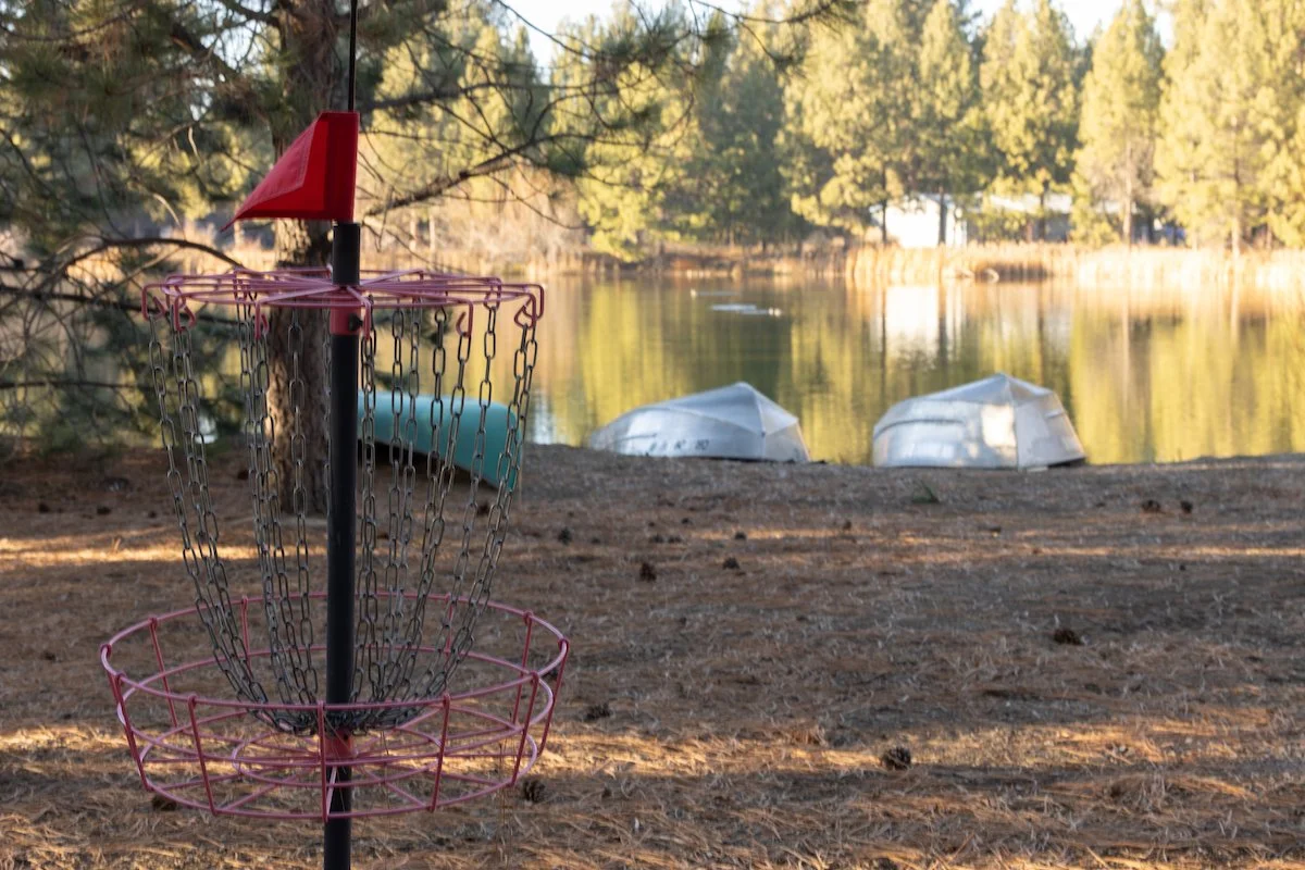A pink and black disc golf basket set up on a dirt ground near a lake with trees in the background. Two small boats are floating on the water.