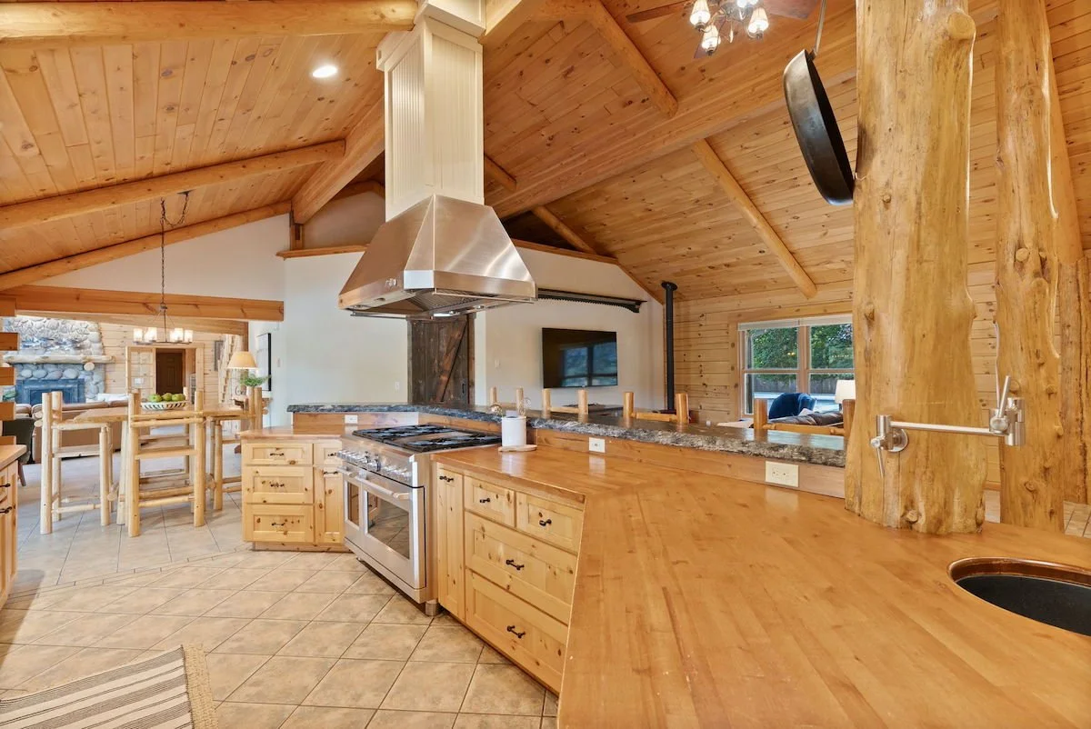 Interior view of a kitchen with wooden cabinetry and vaulted wooden ceiling. There is a stainless steel oven, a granite countertop, a wood support beam, and a living room area in the background.