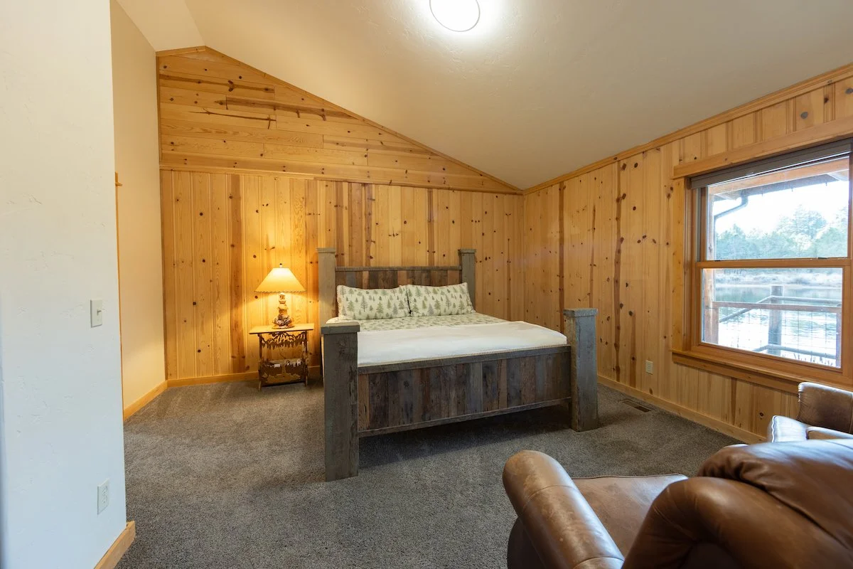 A rustic bedroom with wooden paneling on the walls and ceiling, a large bed with wooden posts, a nightstand with a lamp, and a window showing an outdoor scene.