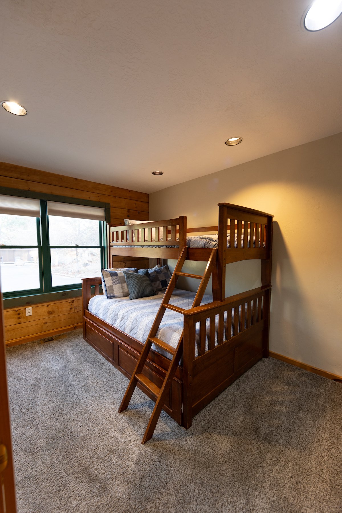 A bedroom with a wooden bunk bed next to a window, with beige walls and carpeted floor.