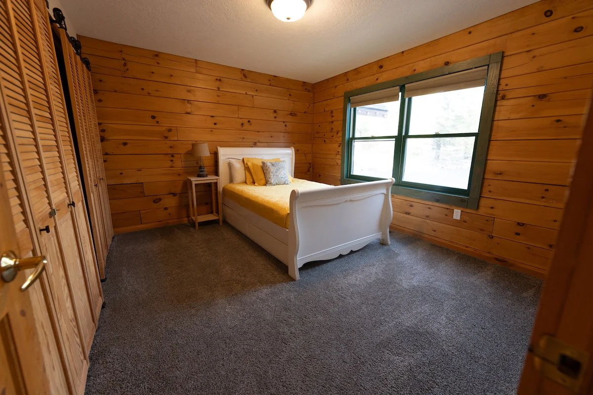 A cozy bedroom with wooden paneled walls, a white bed with yellow bedding and decorative pillows, a bedside table with a lamp, a large window with green trim, and a closet with wooden louvered doors.