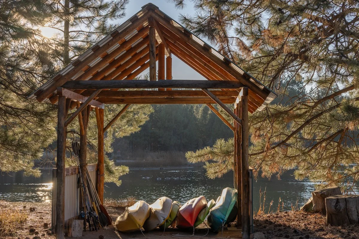 A wooden lakeside shelter with canoes lined up underneath, surrounded by pine trees, overlooking a calm lake during sunset.