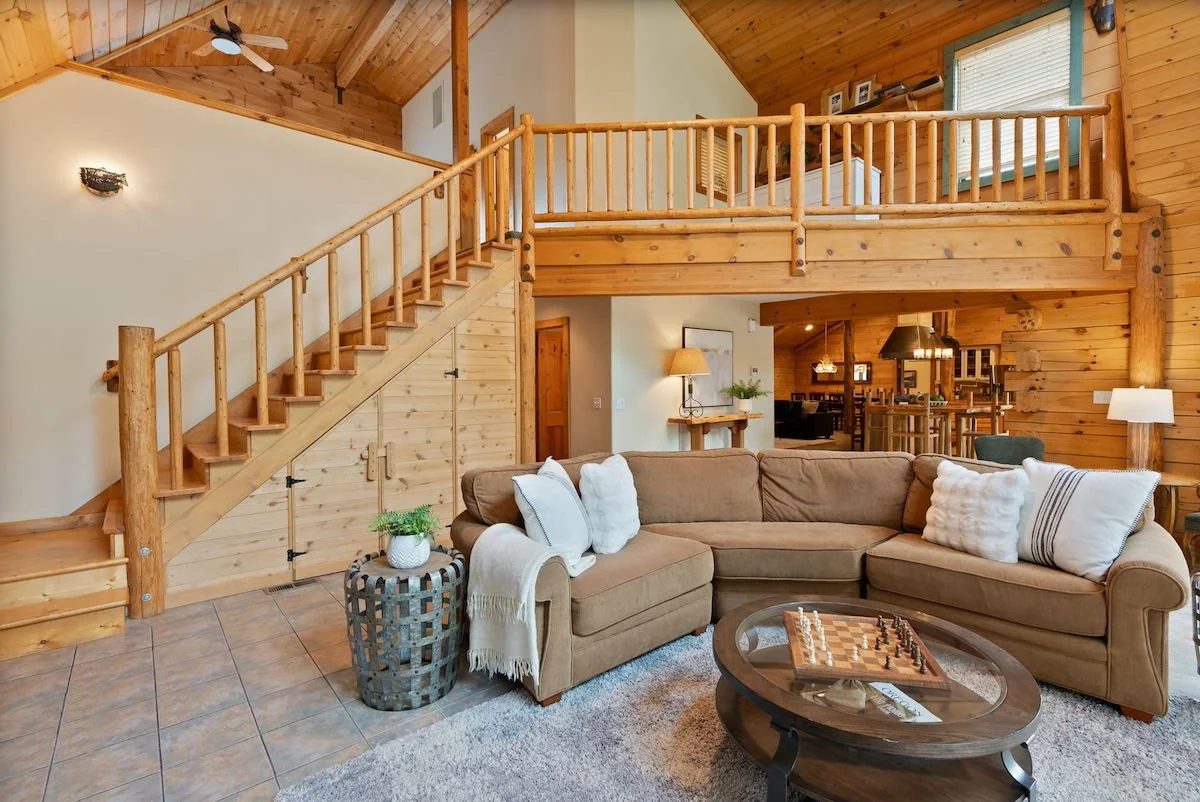 Living room with wooden walls and ceiling, a beige sectional sofa with white and patterned pillows, a round glass-top coffee table with a chessboard, and a potted plant on a basket beside the sofa. A staircase with wooden railing leads to an upper le
