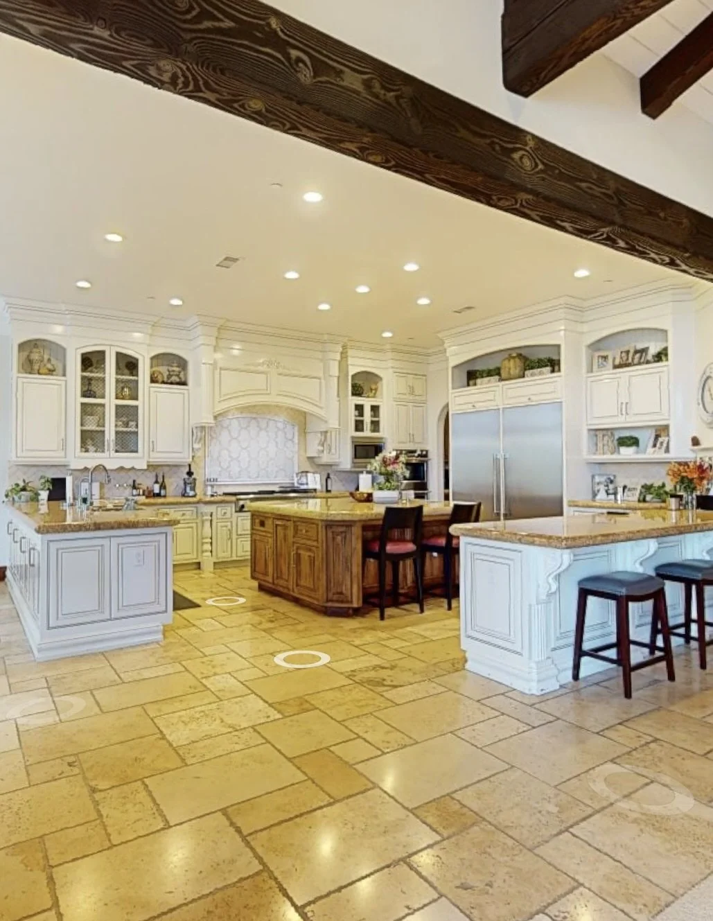 Luxury kitchen interior by Savvy Interiors in Los Angeles, CA, featuring custom white cabinetry, large central island, stone flooring, exposed wood ceiling beams, and high-end appliances in a classic Mediterranean-style home.