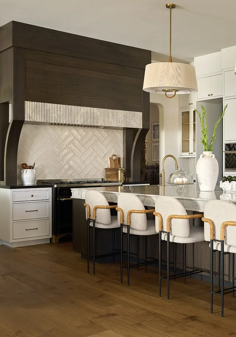 Modern kitchen design by Savvy Interiors in Los Angeles, CA, featuring a dark wood range hood, white herringbone tile backsplash, marble island, brass fixtures, and upholstered counter stools under a warm pendant light.
