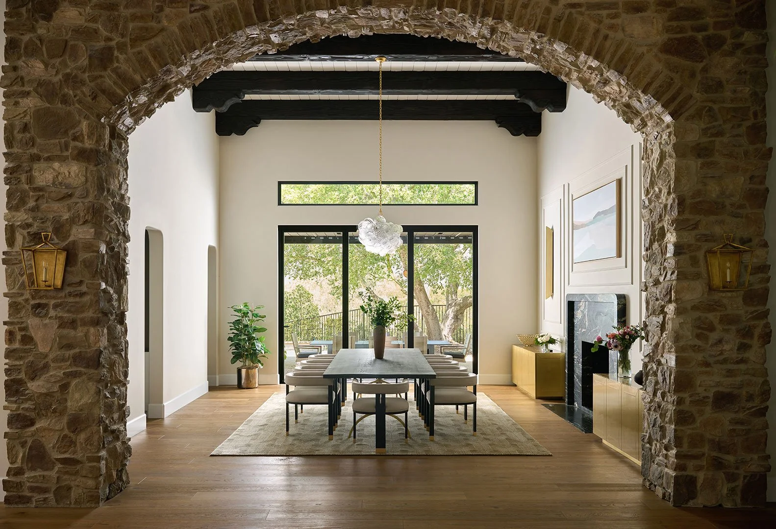 Elegant dining room renovation by Savvy Interiors in Los Angeles, CA, featuring a stone archway, exposed wood ceiling beams, modern chandelier, and a refined indoor-outdoor entertaining space.