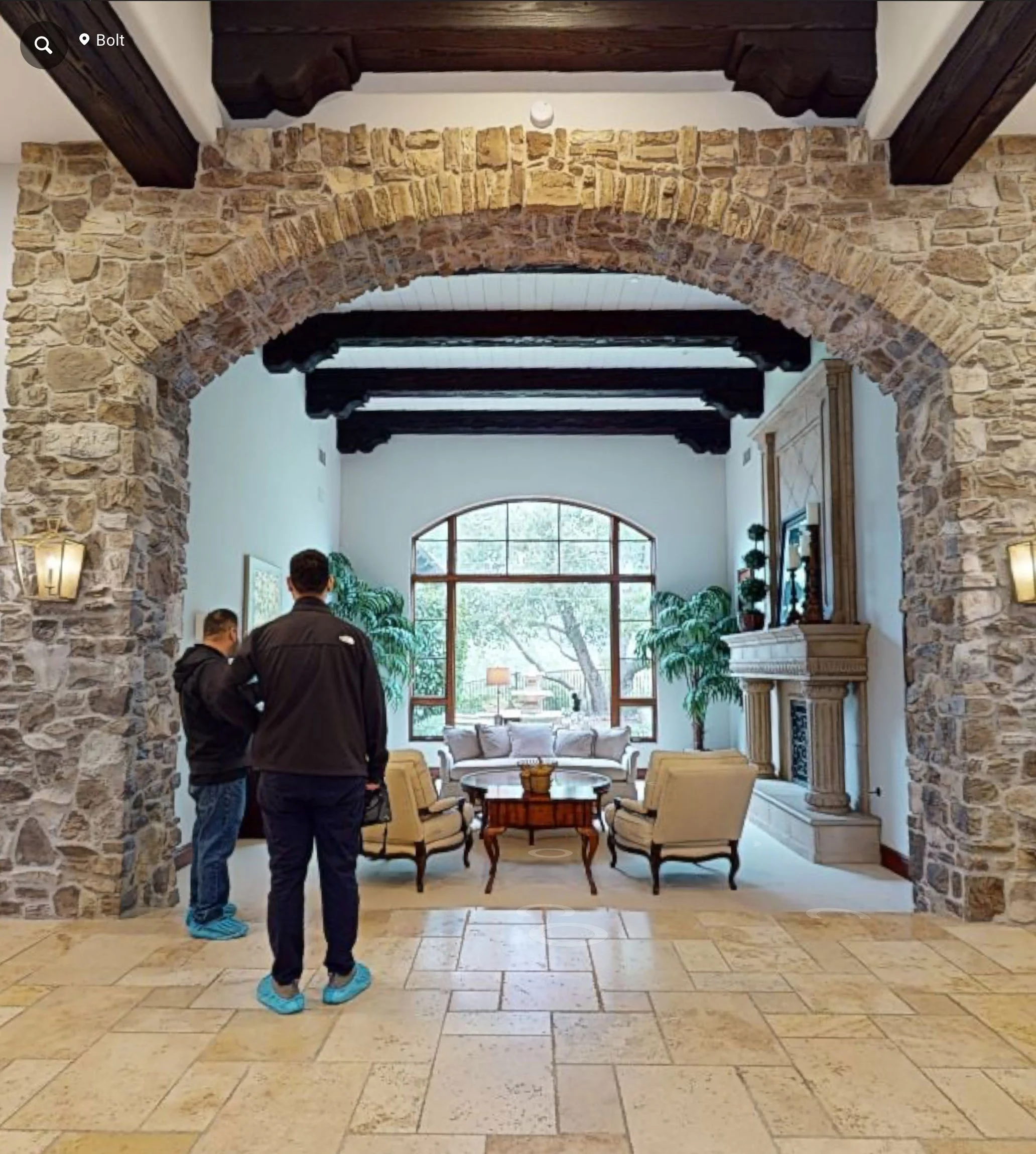 Stone archway framing a luxury living room designed by Savvy Interiors in Los Angeles, CA, featuring exposed wood ceiling beams, limestone floors, classic seating, and a custom fireplace with natural light-filled windows.