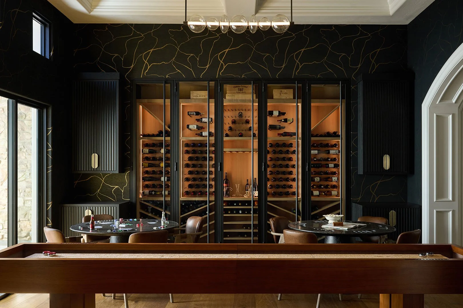 Custom luxury wine room designed by Savvy Interiors in Los Angeles, CA, featuring floor-to-ceiling glass wine cabinets, black accent walls with gold detailing, and an elegant entertaining space.