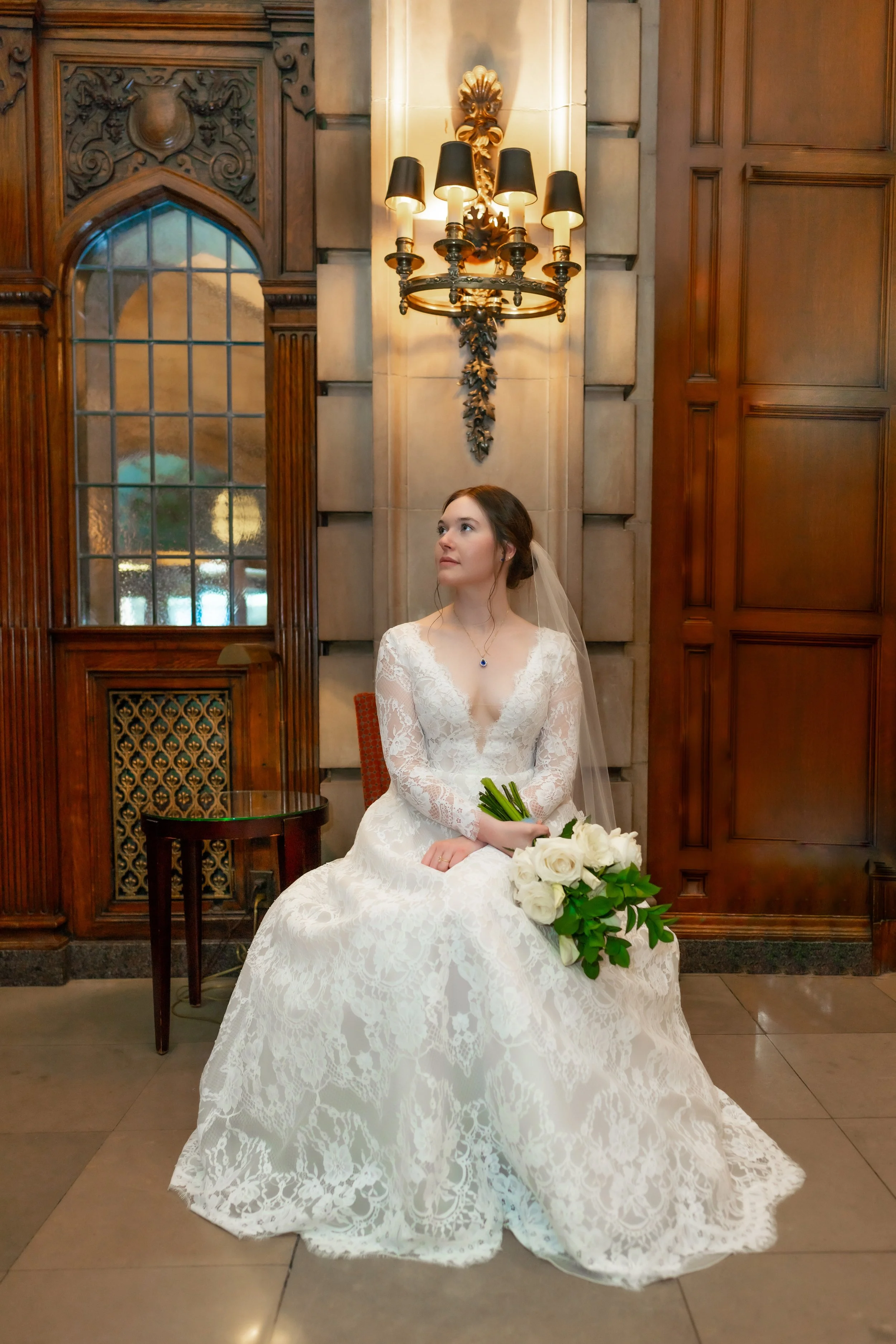 A bride sitting on a chair in an ornate room with wooden paneling, a large window, and a chandelier, holding a bouquet of white roses and looking to her left.