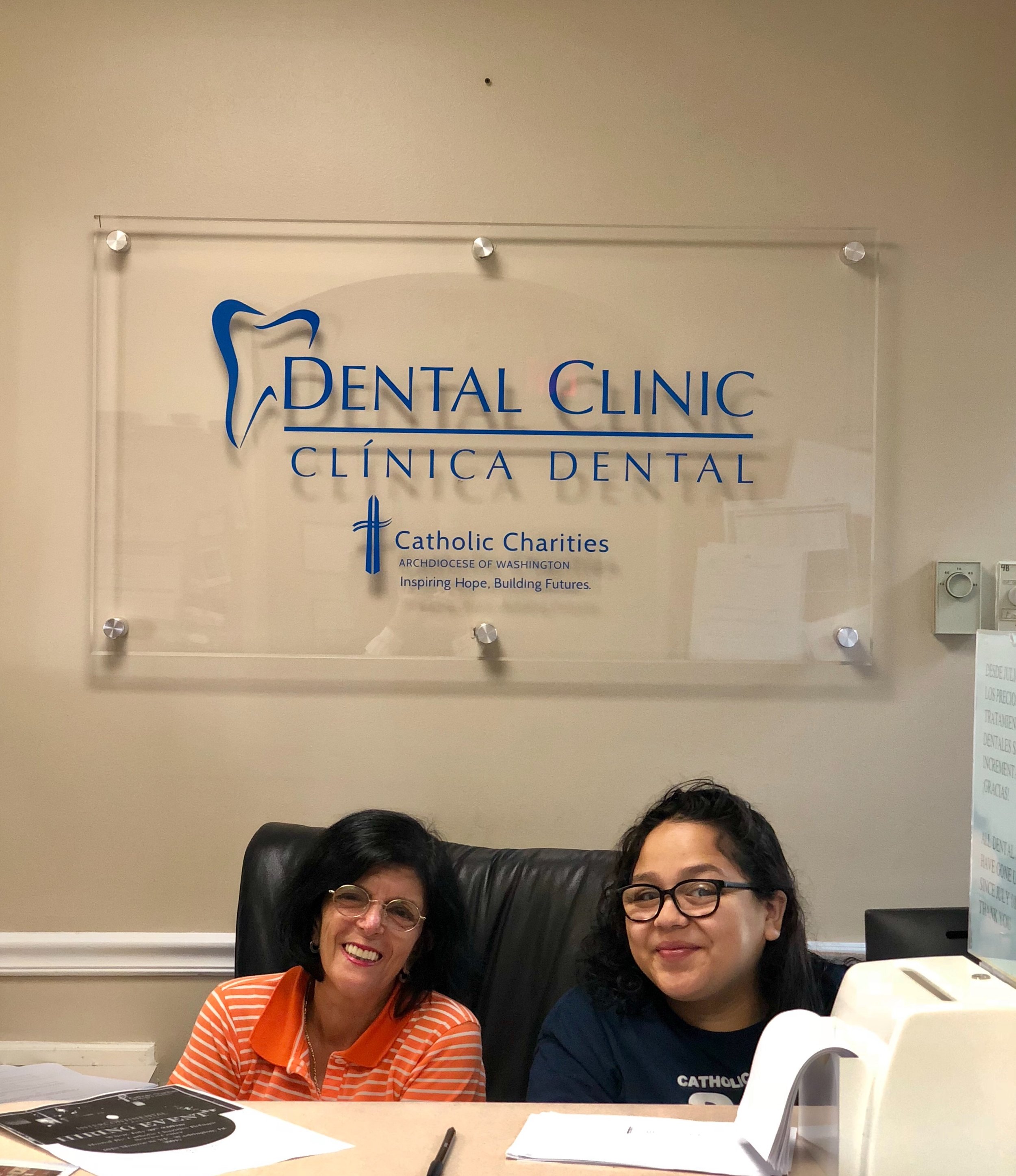 Two smiling women sitting at front desk. A Dental Clinic Catholic Charities sign hangs in the background