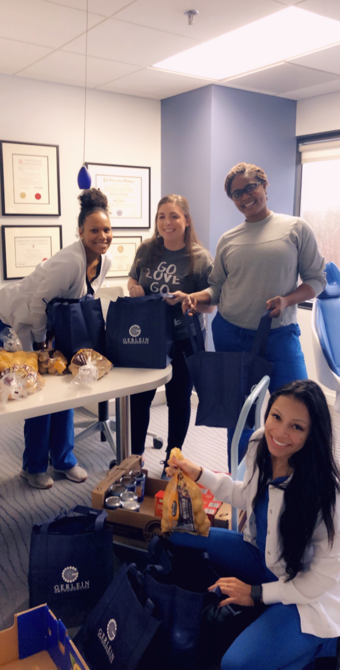 Gerlein team members standing in an office, smiling at the camera holding Gerlein Orthodontics bags. The bags are being filled with thanksgiving food to be dontated. More bags are on the floor