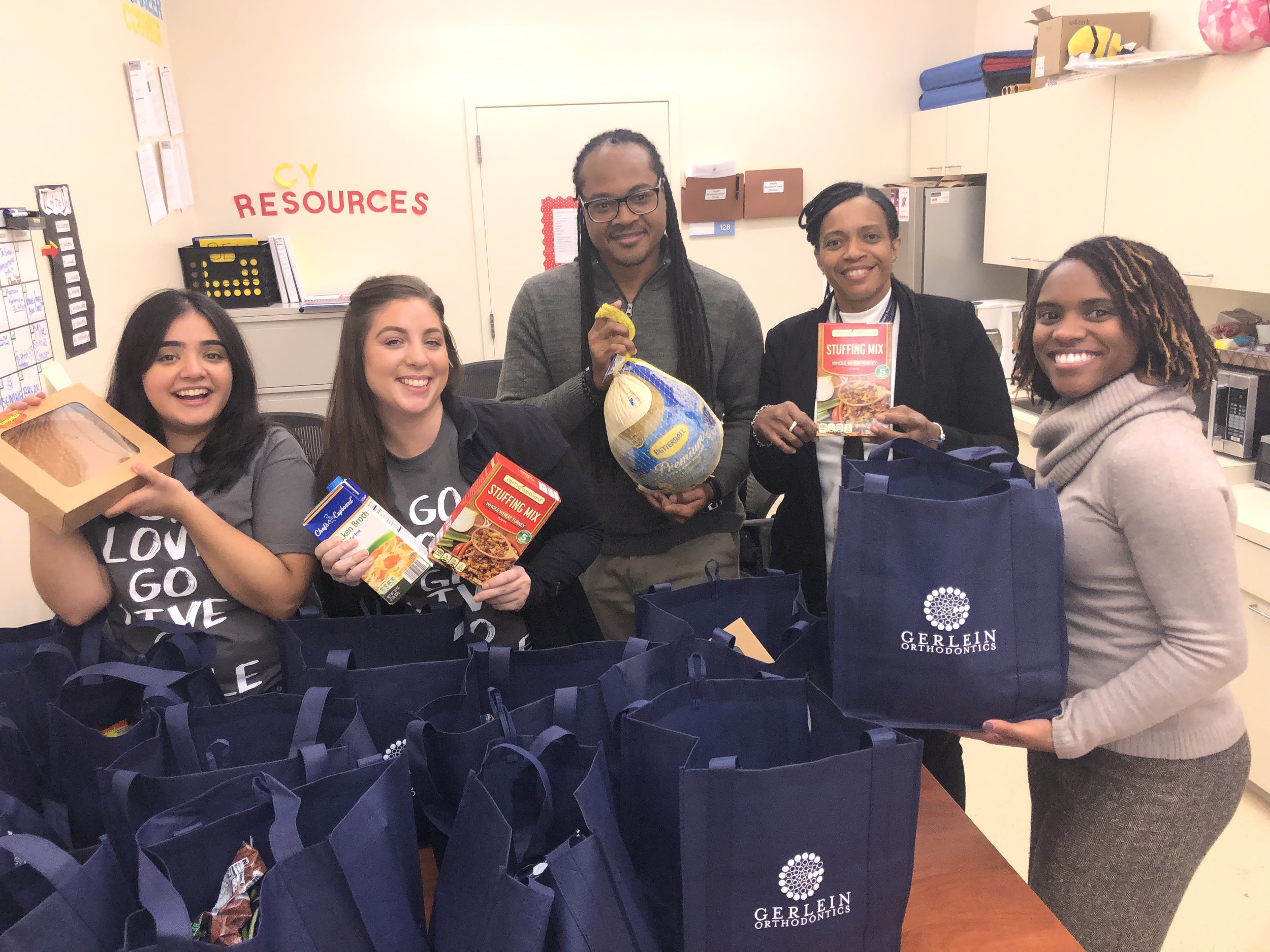 Gerlein staff members stand smiling with members of a donation center. Everyone is holding thanksgiving food items and Gerlein bags.