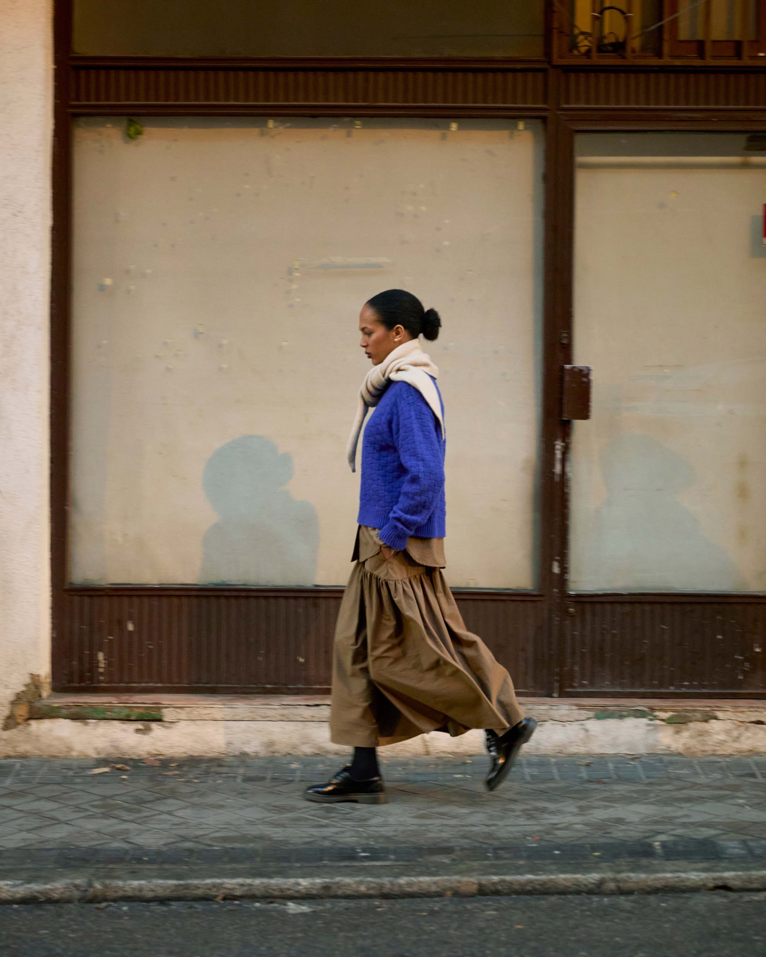 A woman walking on a city sidewalk with her eyes closed, wearing a blue sweater, beige skirt, black shoes, and a scarf, in front of a weathered wall and a closed wooden storefront.