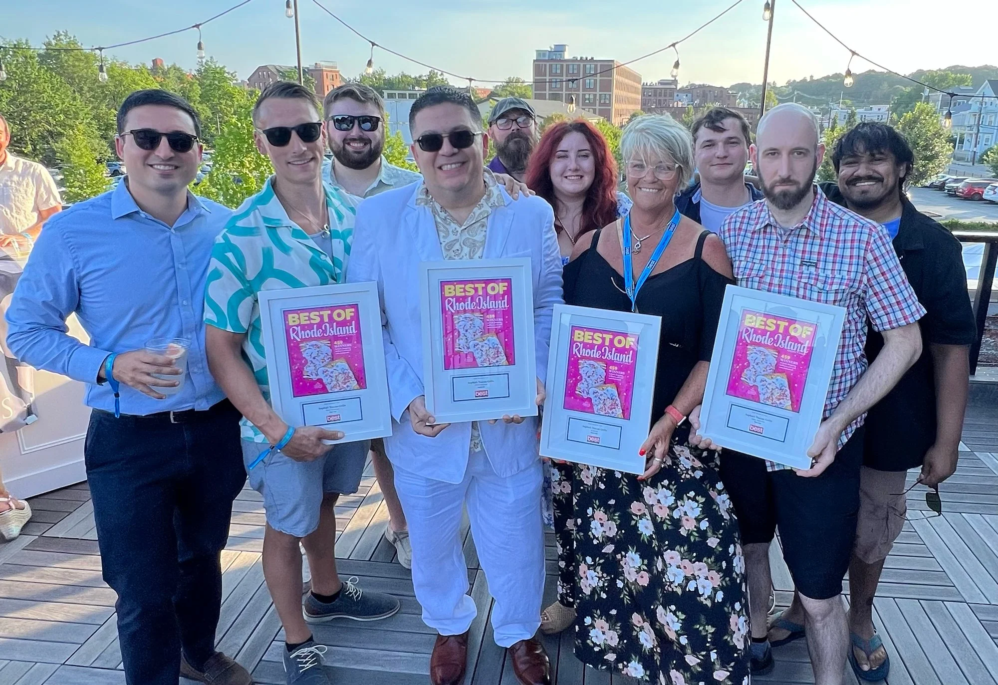Group of eleven people smiling outdoors at sunset, holding framed awards that say "Best of Rhode Island," with trees, buildings, and string lights in the background.