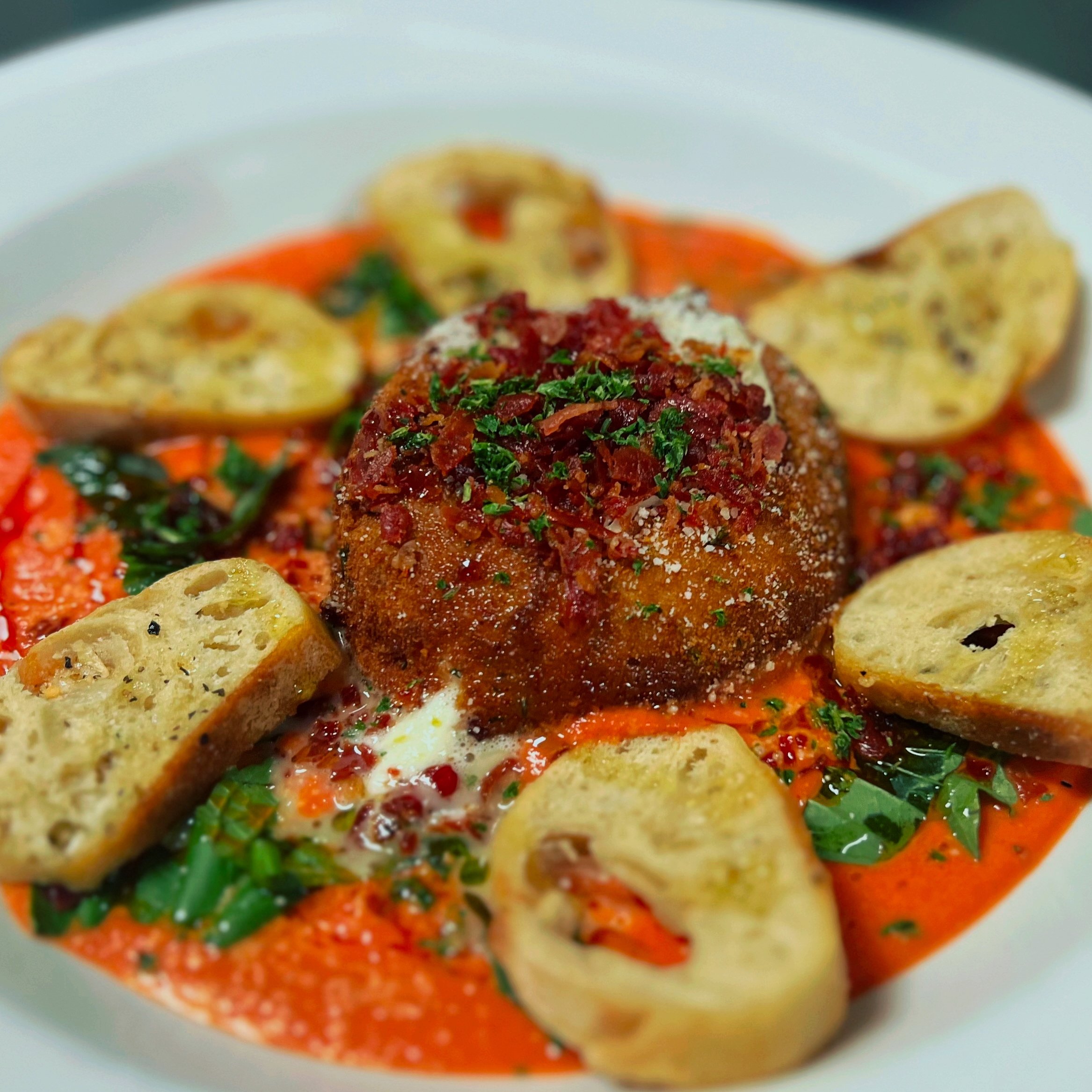 Close-up of a gourmet dish featuring a breaded meatball topped with tomato sauce and chopped herbs, surrounded by toasted baguette slices on a bed of creamy tomato sauce with spinach and red pepper flakes.