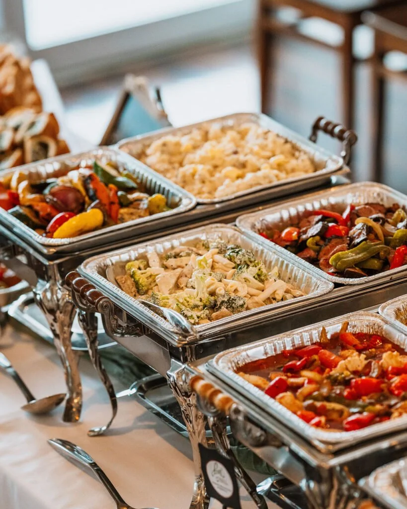 A buffet table with aluminum trays of assorted cooked vegetables, rice, pasta, and stew at a catered event.