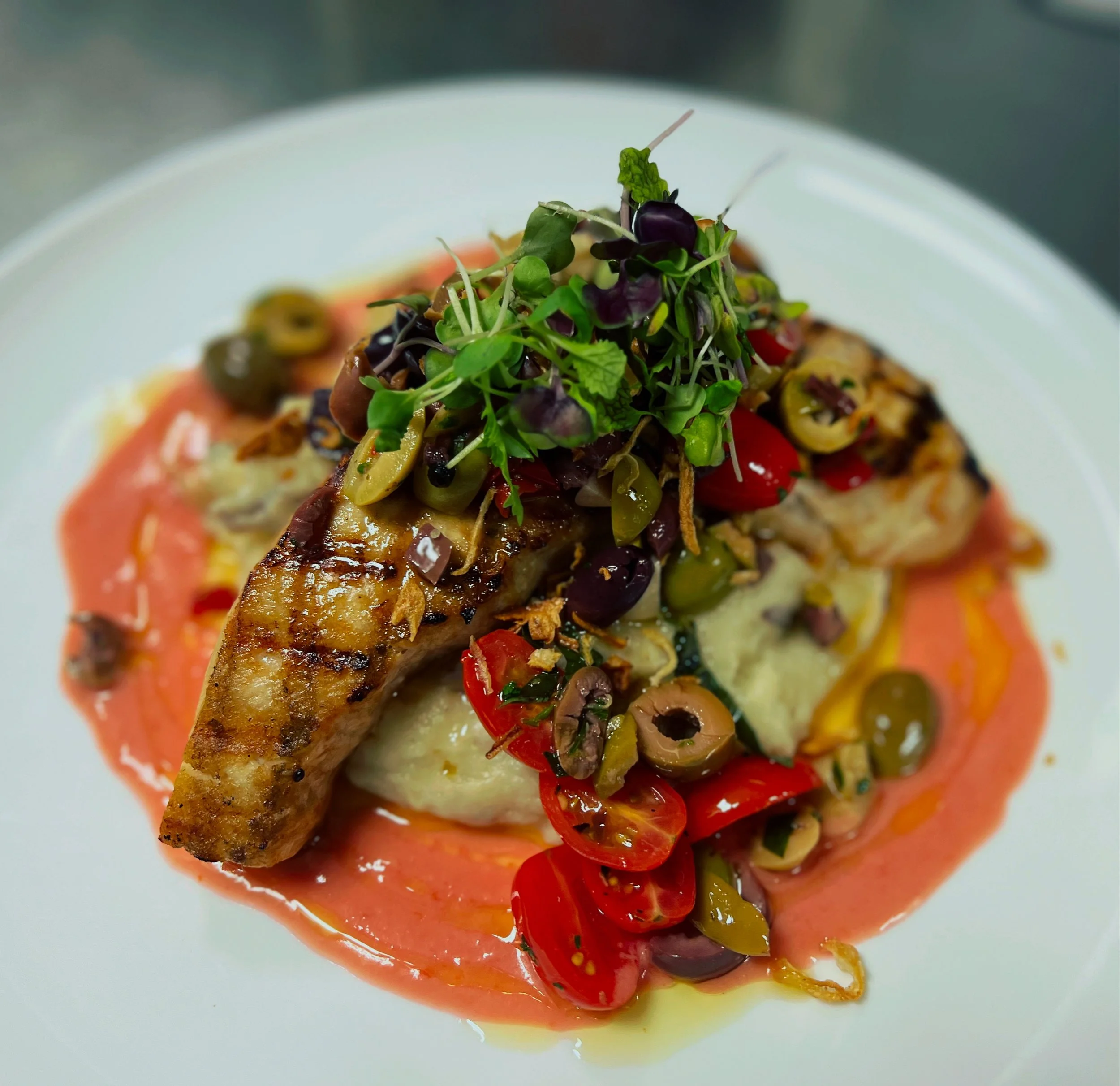 Grilled chicken breast topped with mixed olives, cherry tomatoes, microgreens, and herbs on mashed potatoes with pink sauce on a white plate.