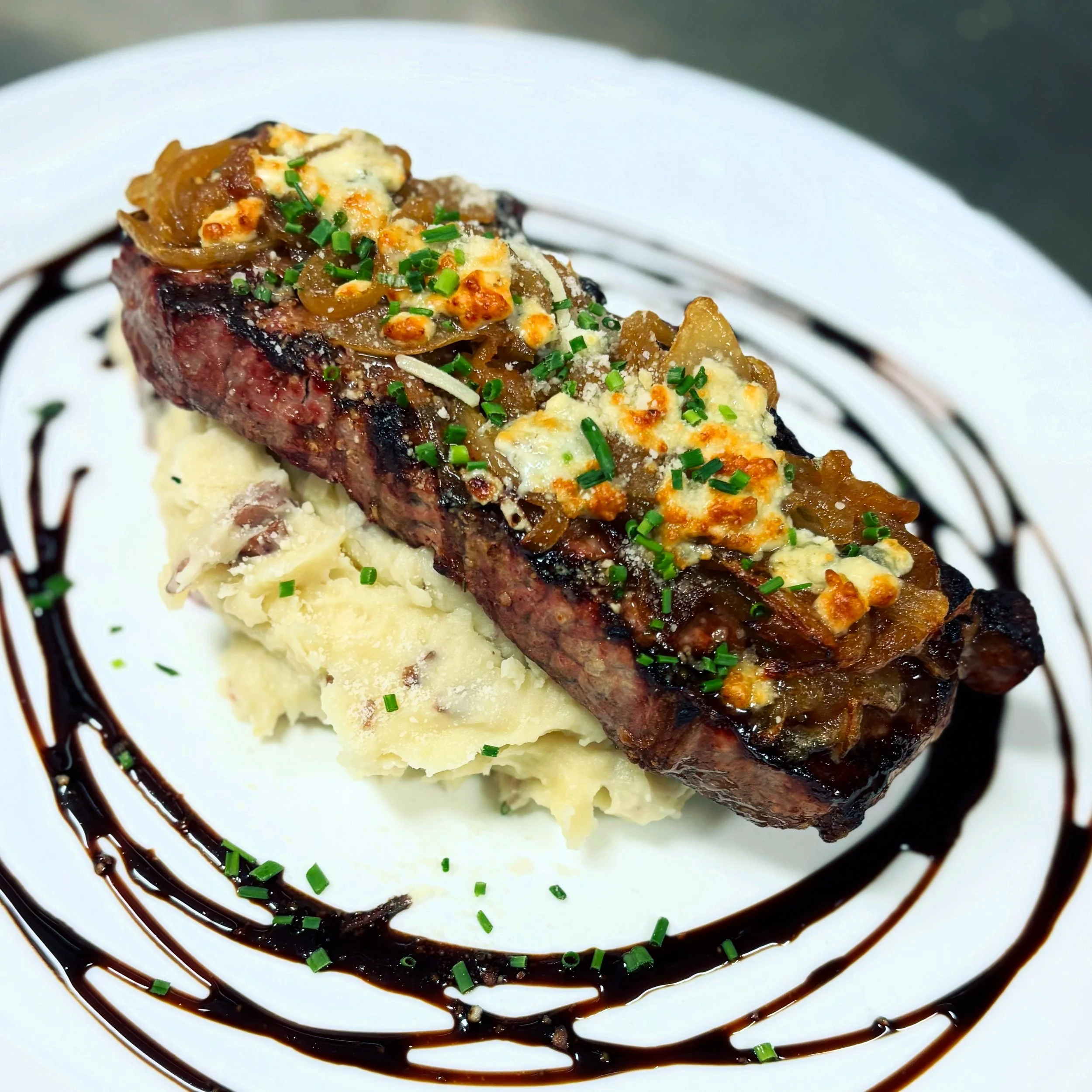 A plated dish with a grilled beef steak topped with caramelized onions, crumbled cheese, and chopped chives, served over mashed potatoes with a decorative drizzle of balsamic glaze on the plate.