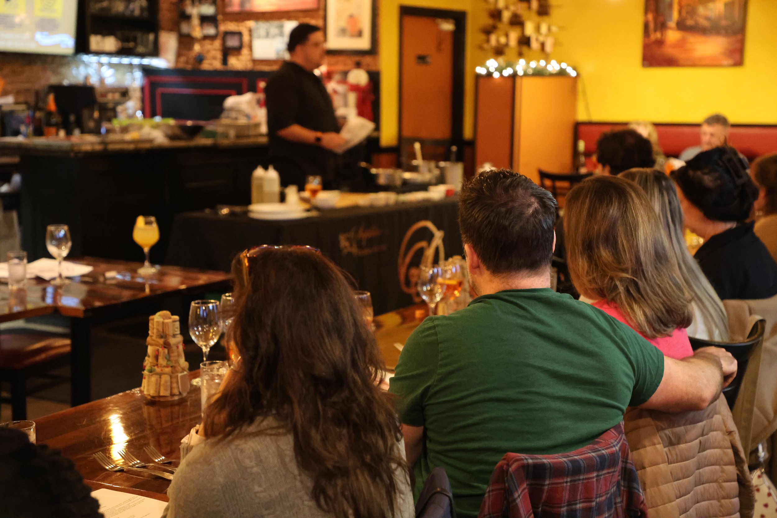 People sitting at tables watching a chef prepare food in a restaurant with warm lighting and yellow walls.