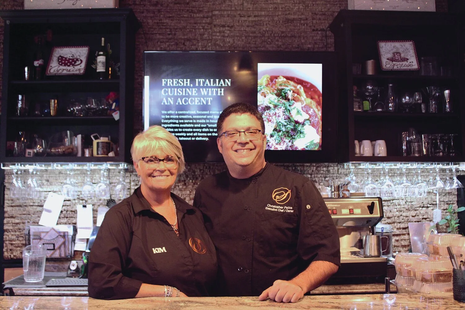 Two smiling people, a woman and a man, standing behind the counter of a restaurant kitchen, with a digital menu display in the background.