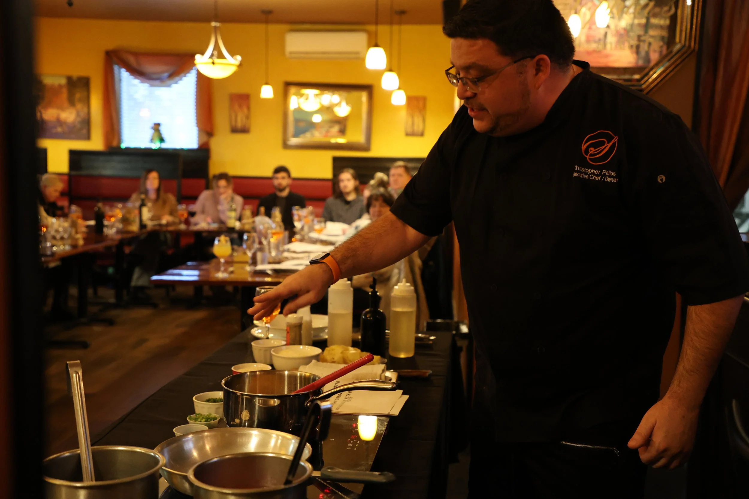 Chef preparing dishes at a restaurant with diners seated at tables in the background.