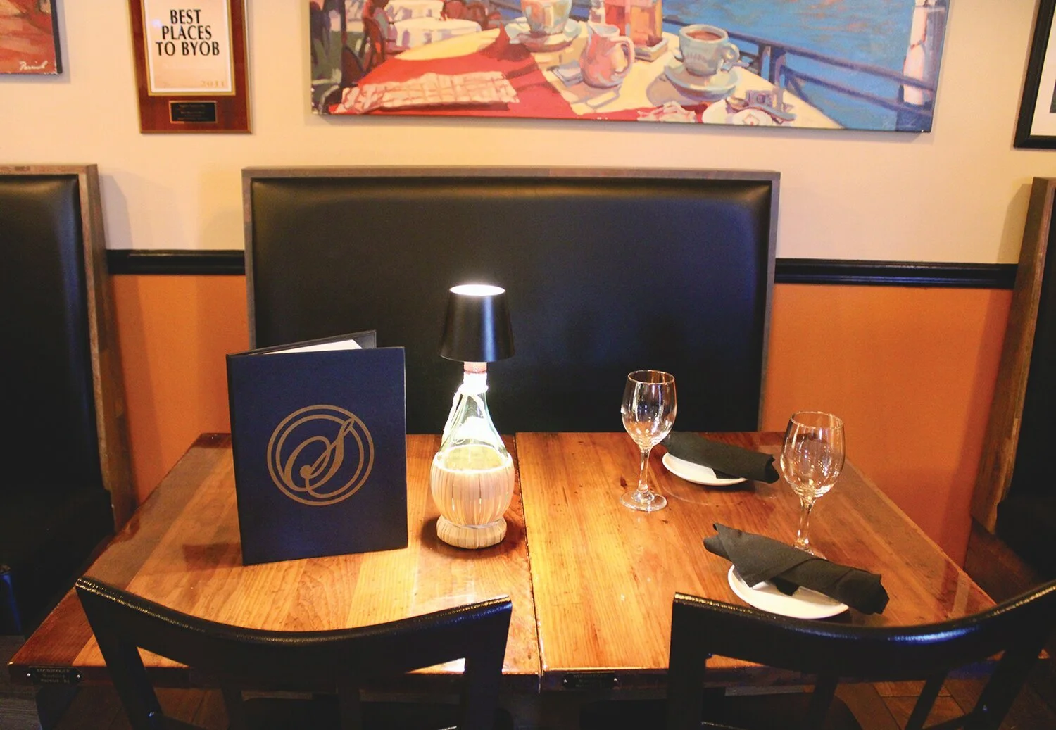 A dining table set with two plates with black napkins, two wine glasses, a menu, a small lamp, and a bottle of water in a restaurant with artwork and a framed sign on the wall.