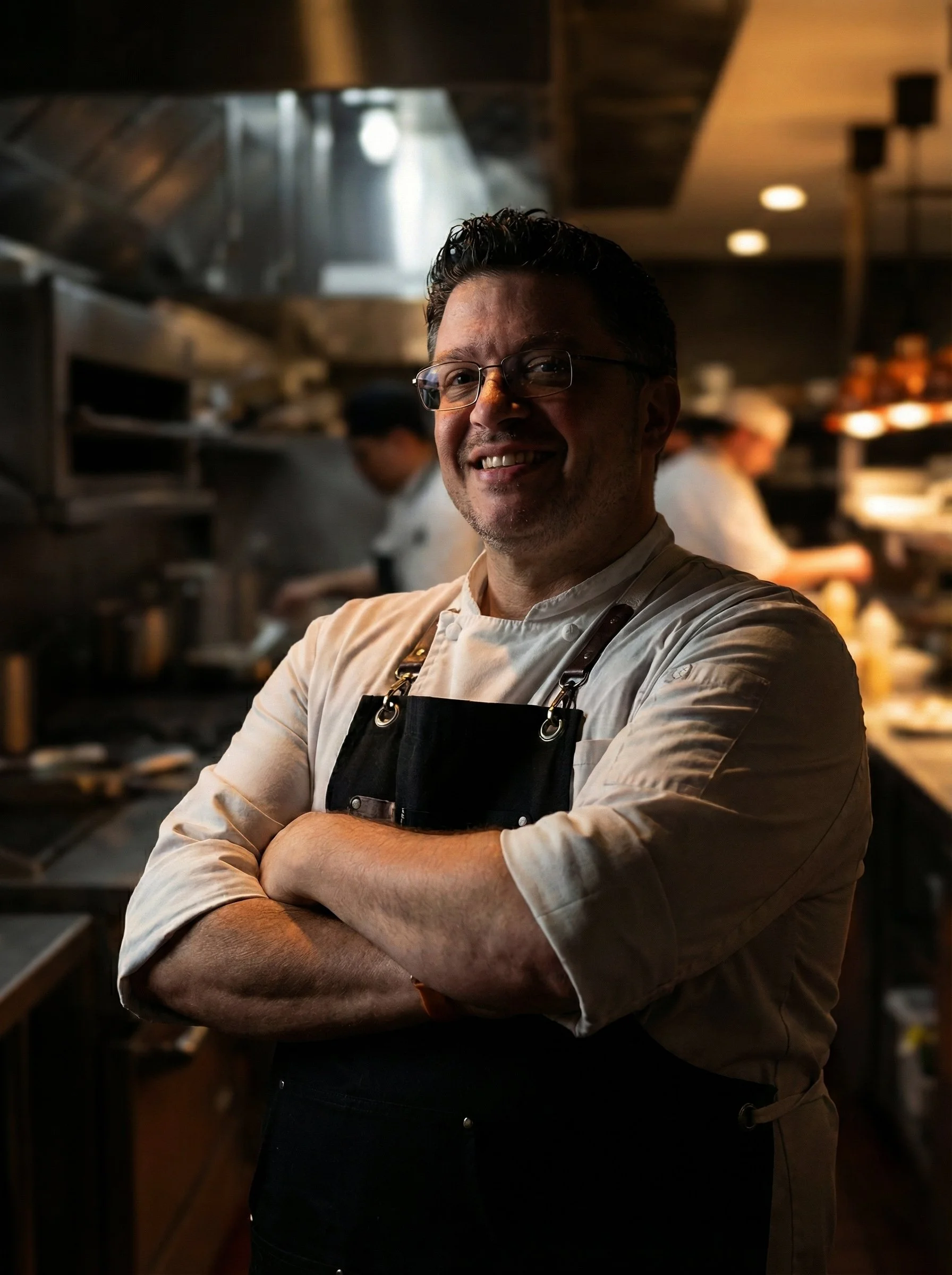 A smiling chef with glasses and short dark hair, wearing a white chef's coat and black apron, standing with arms crossed in a professional kitchen.
