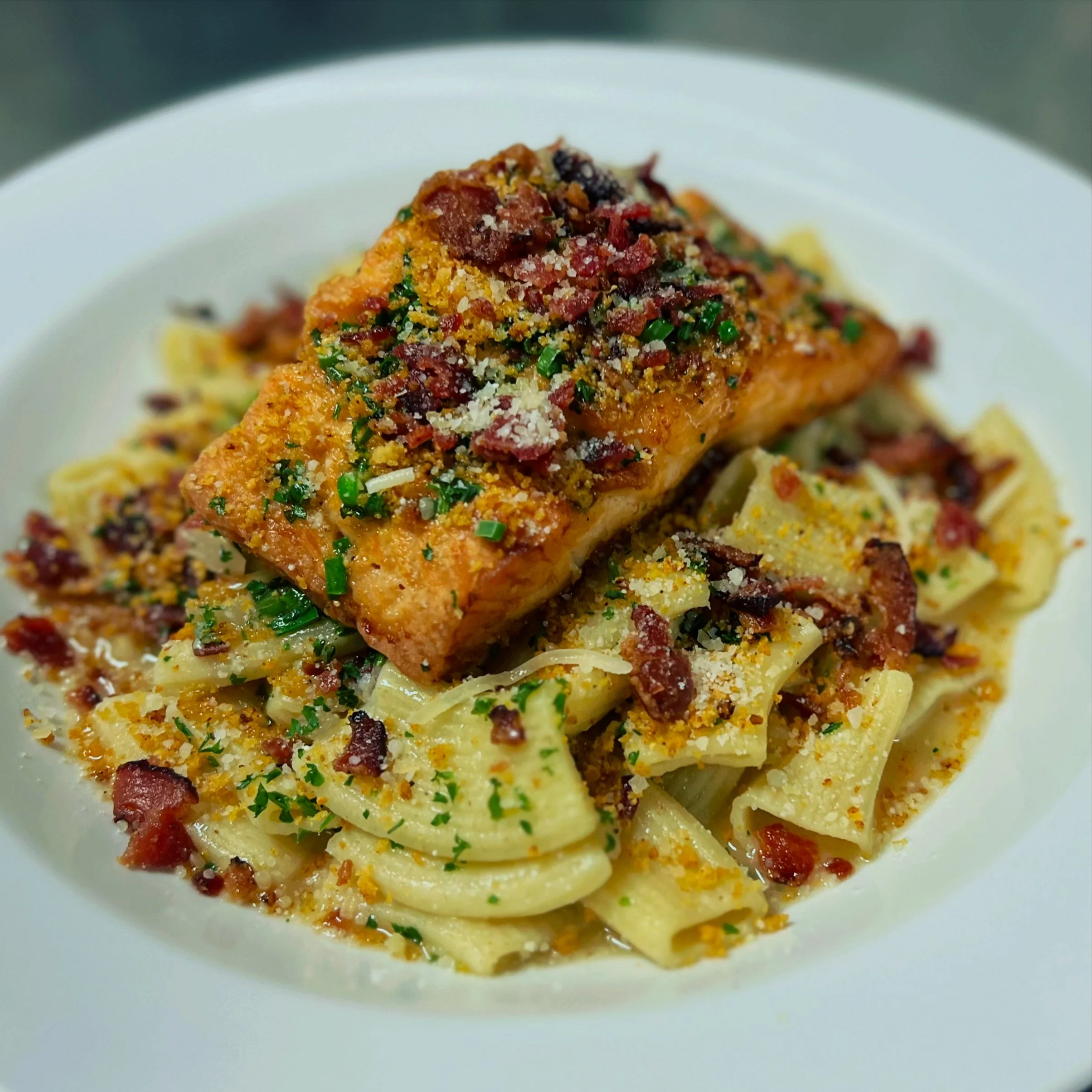 Close-up of a plate with pasta, topped with a grilled salmon fillet garnished with chopped herbs, bacon bits, and grated cheese.
