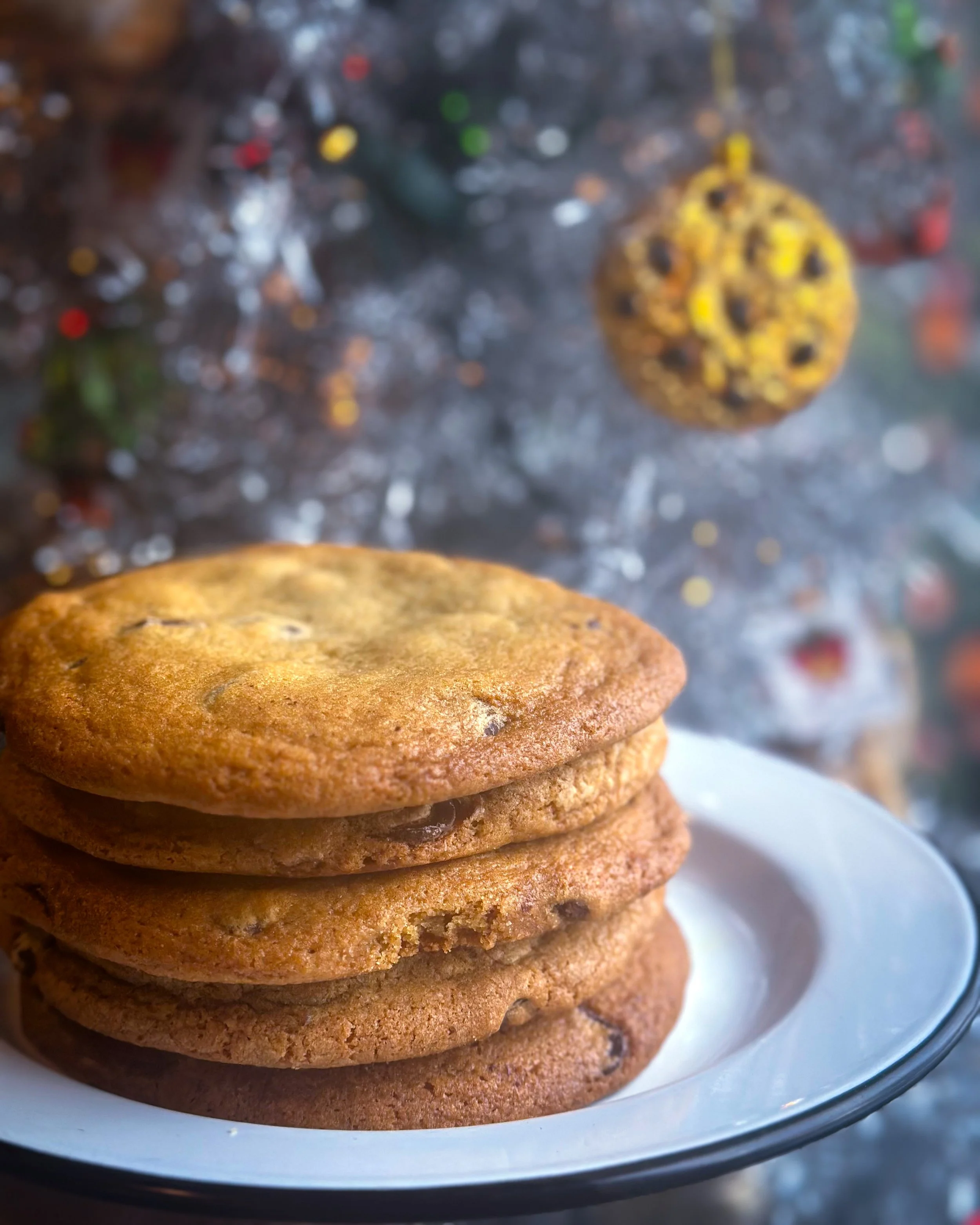 Chocolate chip cookies on a white round plate in front of a silver tinsel tree.
