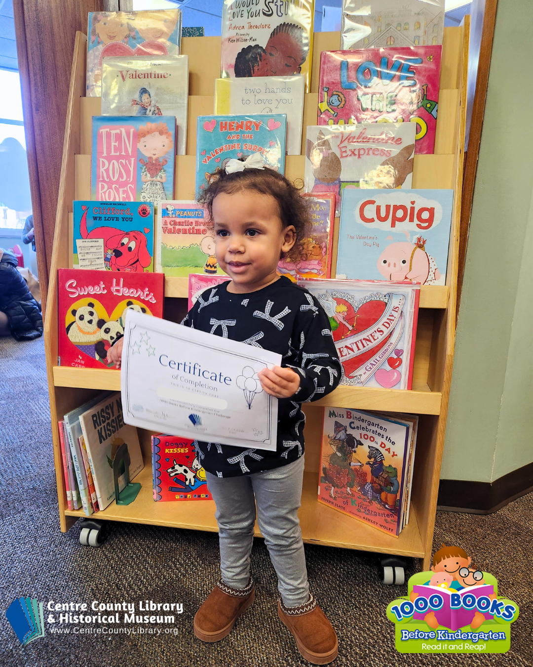 A child holds up their Certificate of Completion in front of a stack of books.